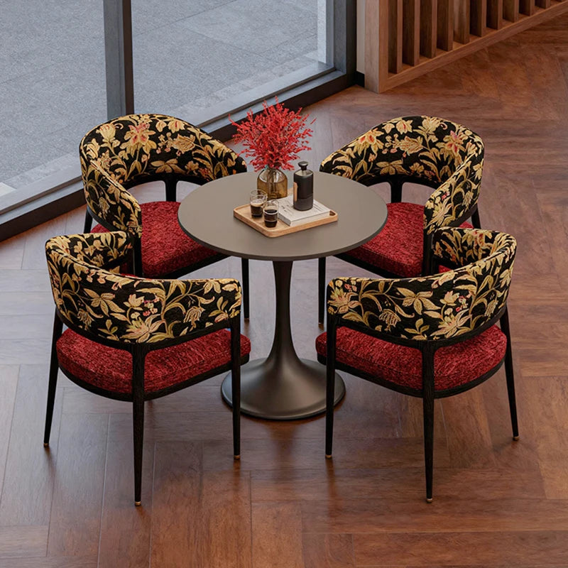 Four ornate chairs with red cushions around a round table in a room with large windows.